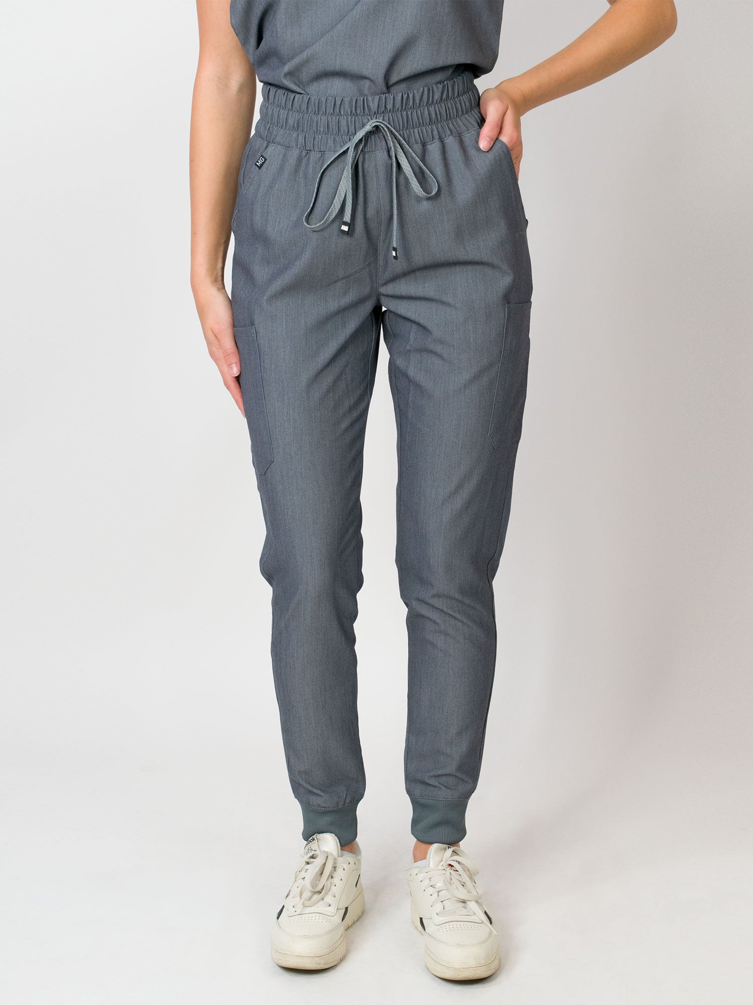 A woman standing in a three-quarter pose wearing gray mid-rise slim jogger scrub pants with an elastic waistband and drawstring, with multiple pockets.