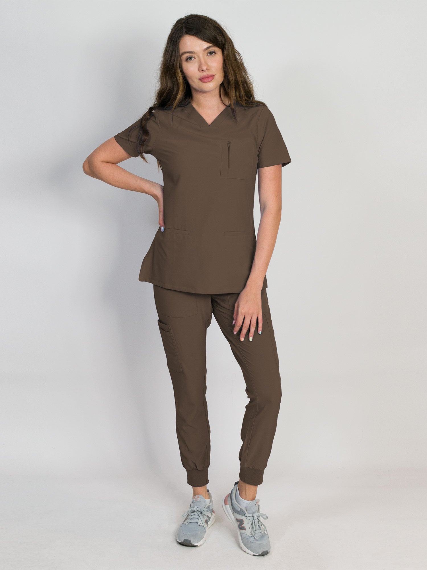 A woman modeling an espresso color scrub set with V-neck mitered neck top and matching jogger pants with multiple pockets, designed for medical professionals.
