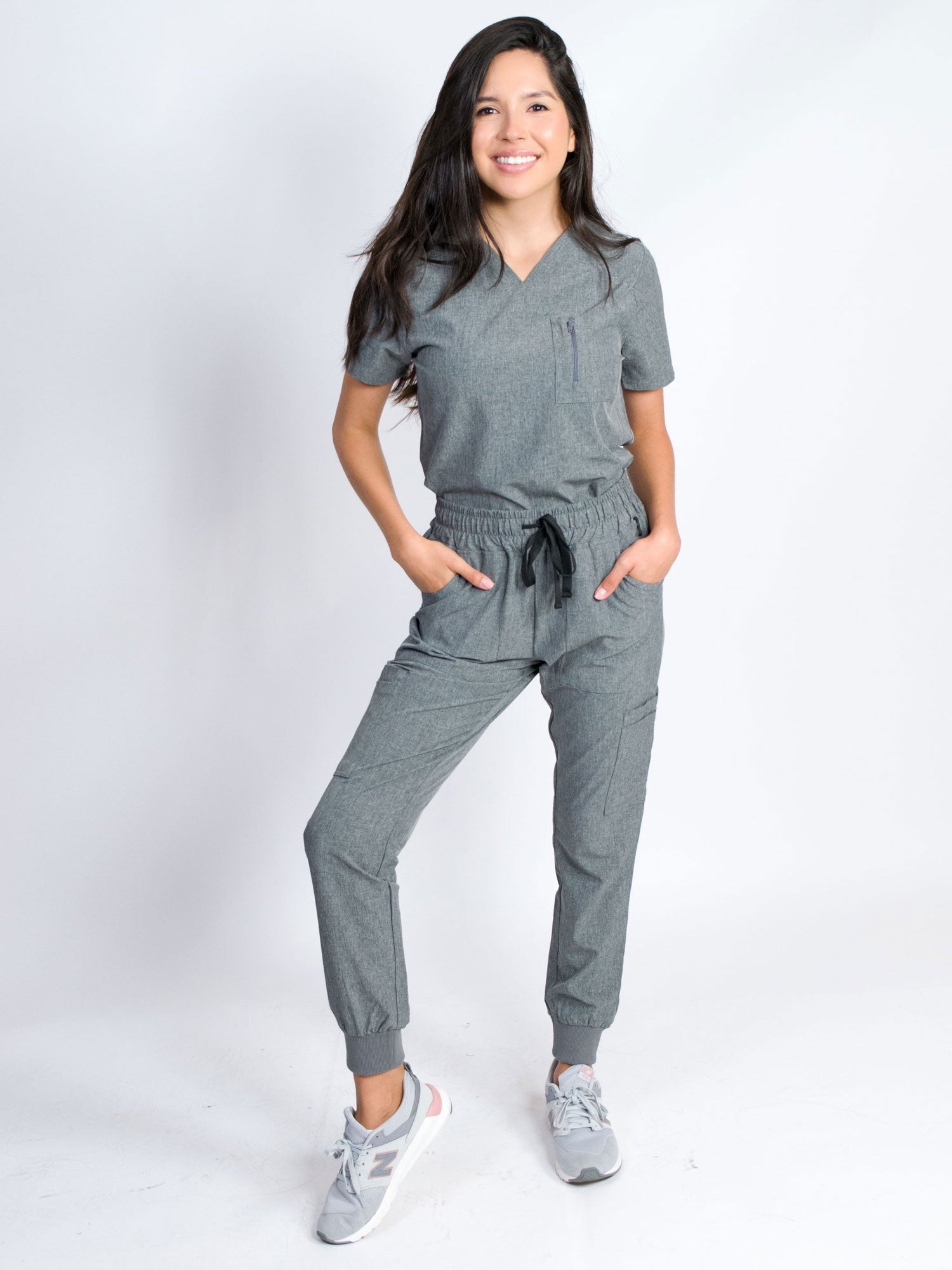 A woman modeling a grey V-neck mitered neck top and matching jogger pants with multiple pockets, designed for medical professionals.