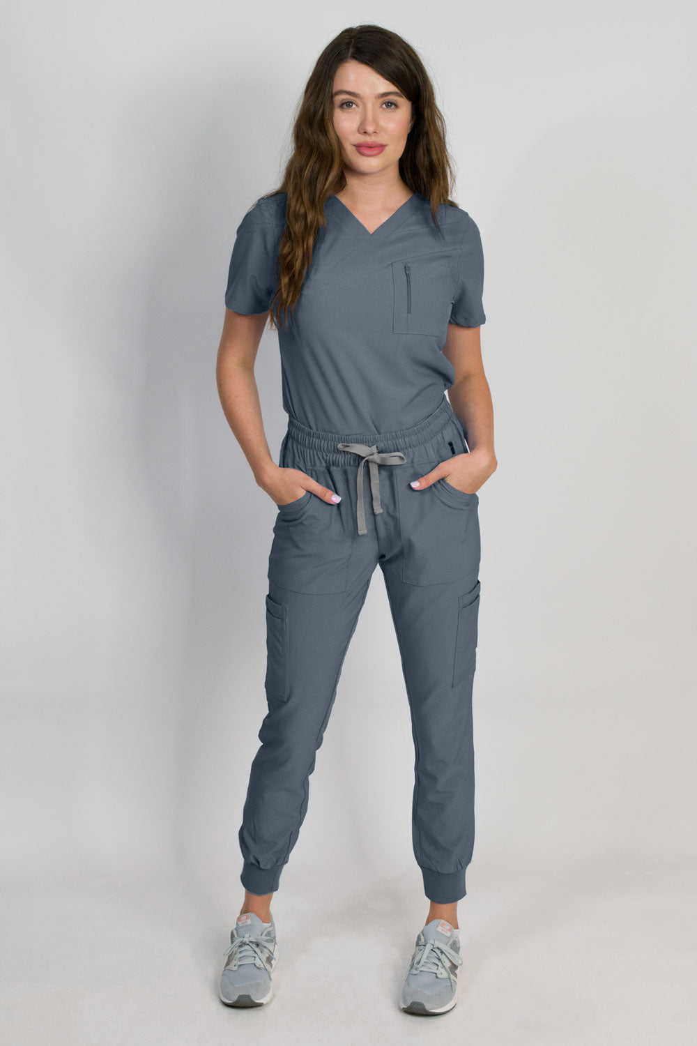 A woman modeling a titanium grey scrub set with V-neck mitered neck top and matching jogger pants with multiple pockets, designed for medical professionals.