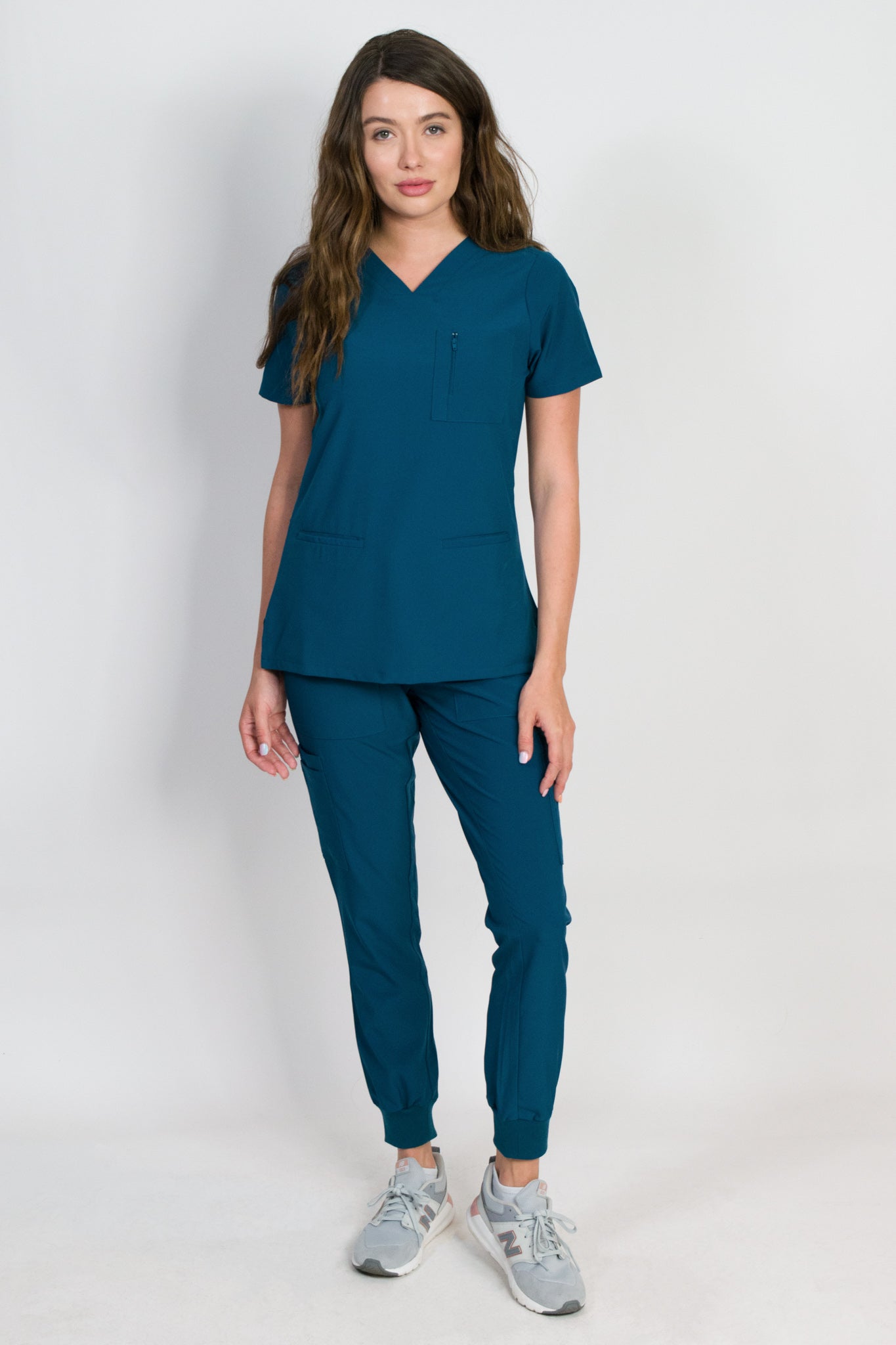 A female model wearing a caribbean green scrub set with mitered neck zip chest pocket top and matching jogger pants with multiple pockets, designed for medical professionals.