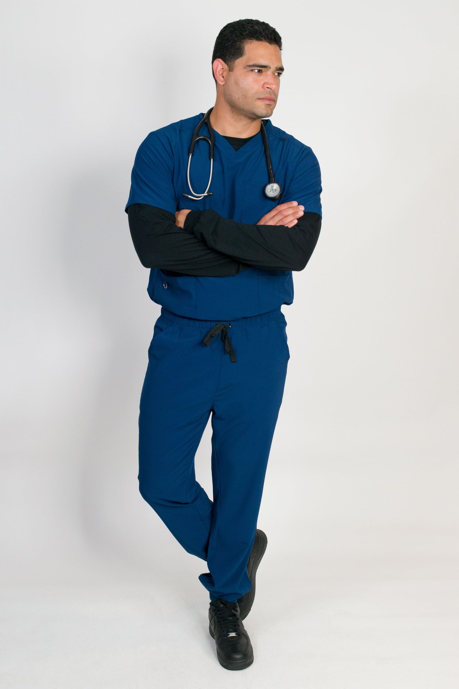 A man wearing a blue V-neck scrub set with 4 pockets and rib knit cuffs. The set includes a top with a chest patch pocket and bottom hem pockets, along with matching pants with inset trouser pockets and back welt pockets.