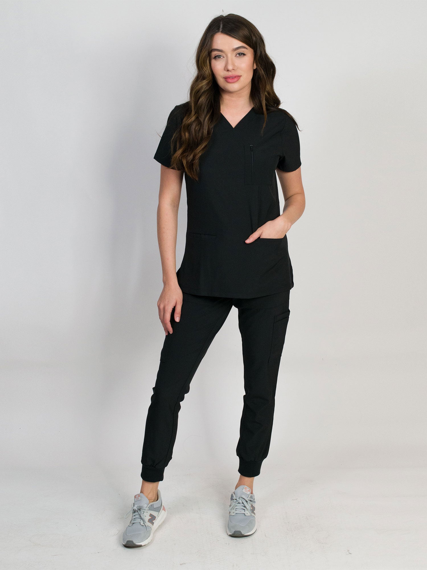 A woman modeling a scrub set in black with V-neck mitered neck top and matching jogger pants with multiple pockets, designed for medical professionals.