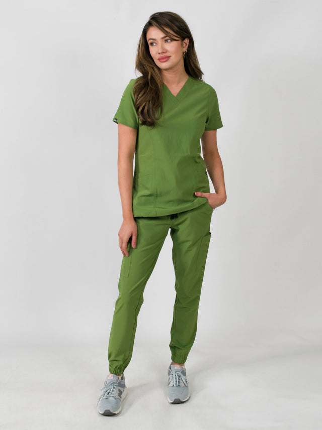 Woman modeling a women's medical scrub set with a V-neck top and jogger pants in avocado green color, featuring multiple pockets.