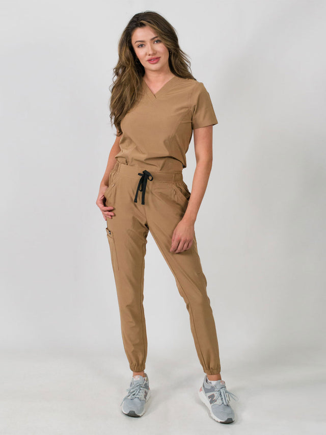 Woman modeling a women's medical scrub set with a V-neck top and jogger pants in brown color, featuring multiple pockets.