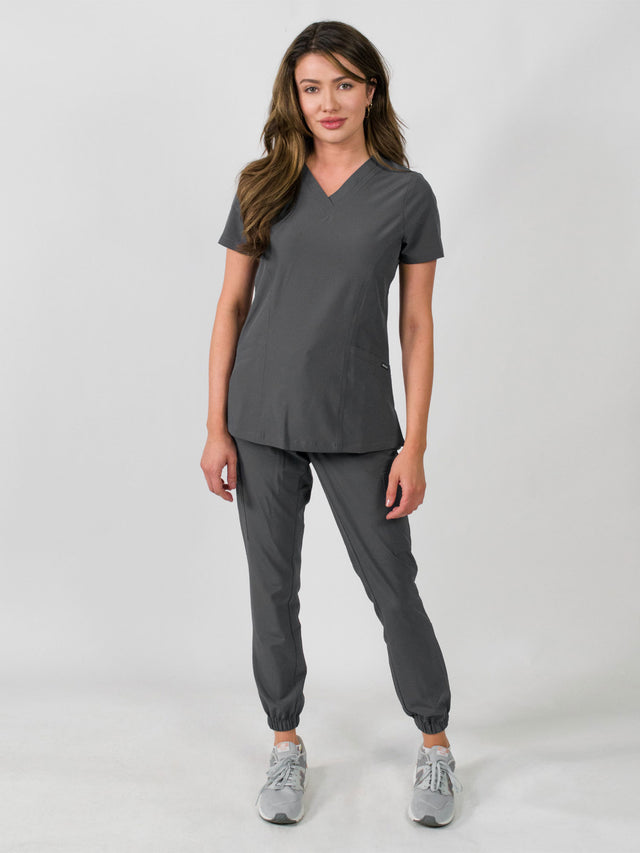 Woman modeling a women's medical scrub set with a V-neck top and jogger pants in grey color, featuring multiple pockets.