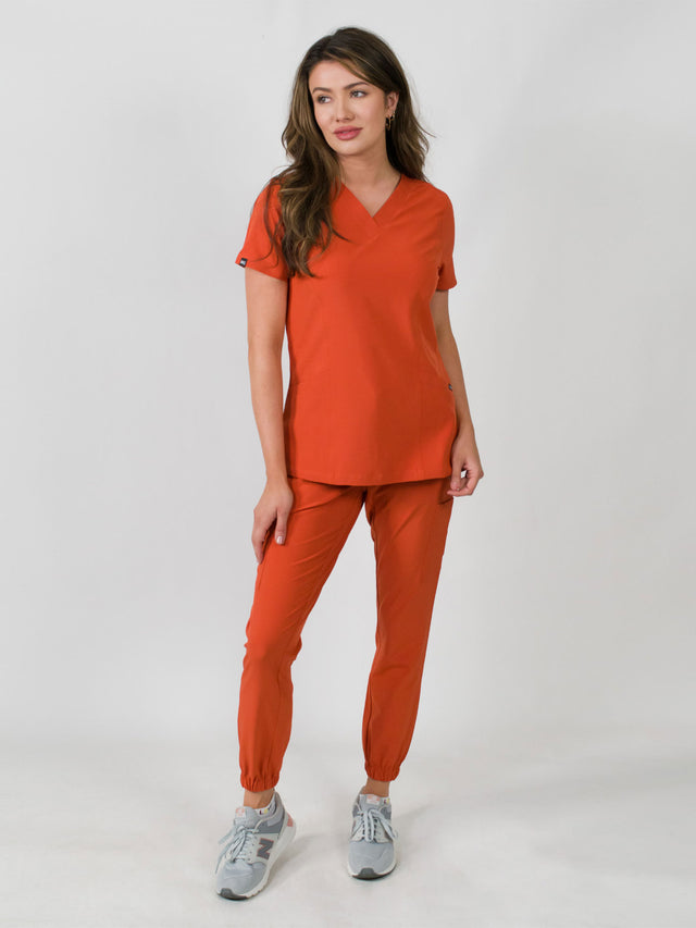 Woman modeling a orange medical scrub set with a V-neck top and jogger pants, featuring multiple pockets.