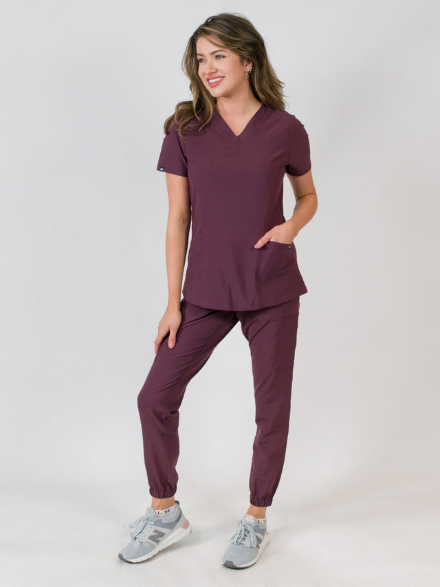 Woman modeling a women's medical scrub set with a V-neck top and jogger pants in purple color, featuring multiple pockets.