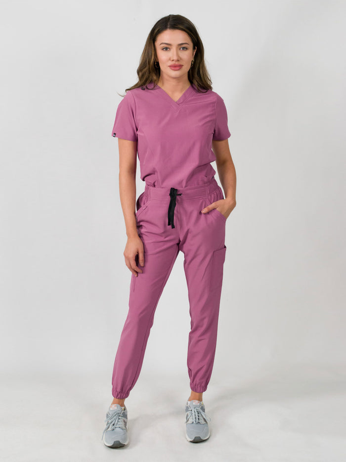 Woman modeling a women's medical scrub set with a V-neck top and jogger pants in rose color, featuring multiple pockets.