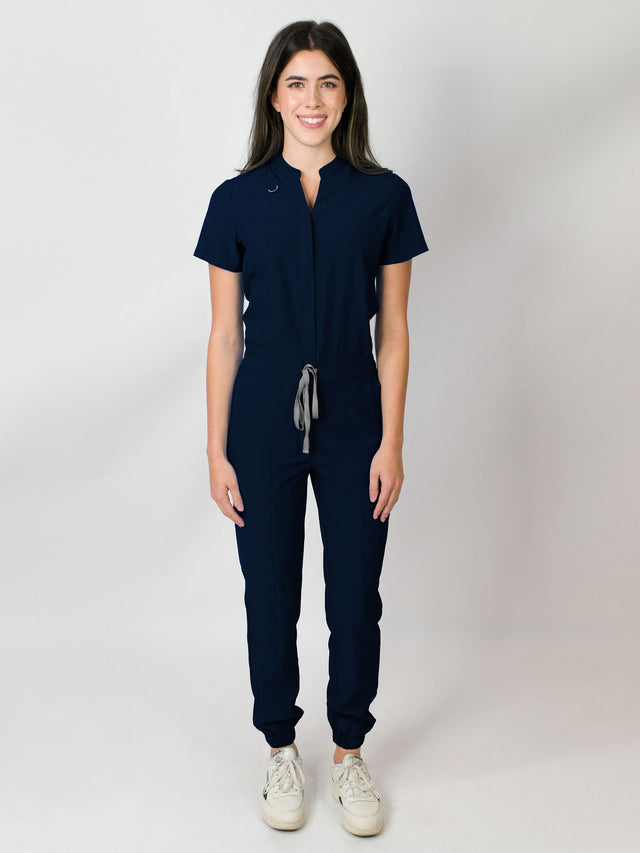 Navy blue scrub jumpsuit with a mandarin collar, short sleeves, and elastic jogger cuffs. The jumpsuit has multiple pockets and a zipper front.