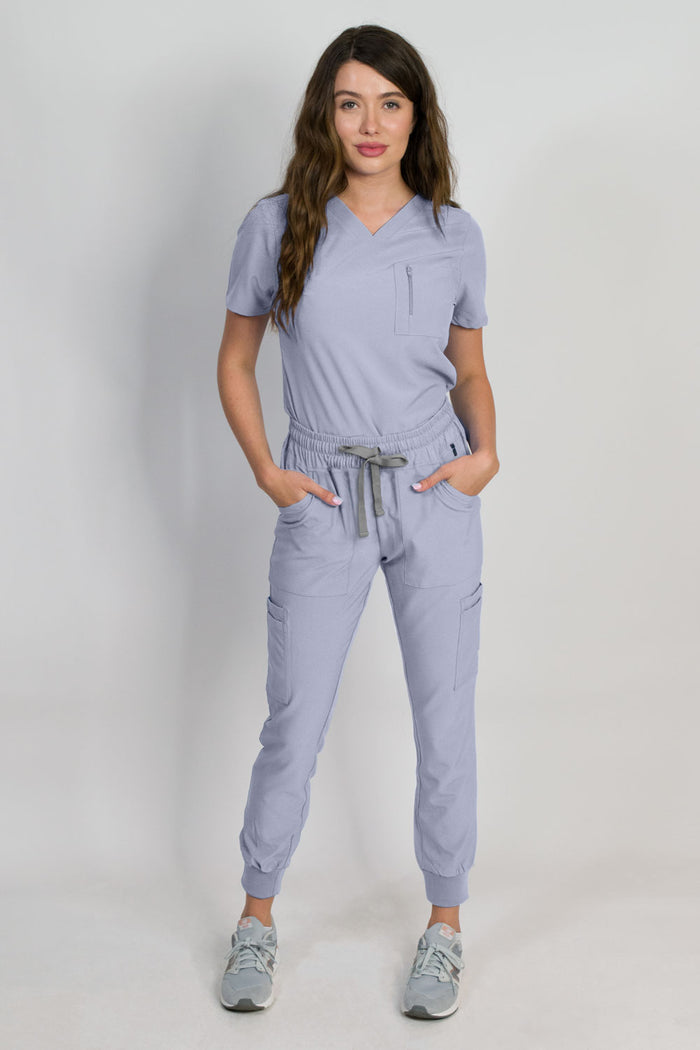 A female model wearing a glacial blue scrub set with mitered neck zip chest pocket top and matching jogger pants with multiple pockets, designed for medical professionals.