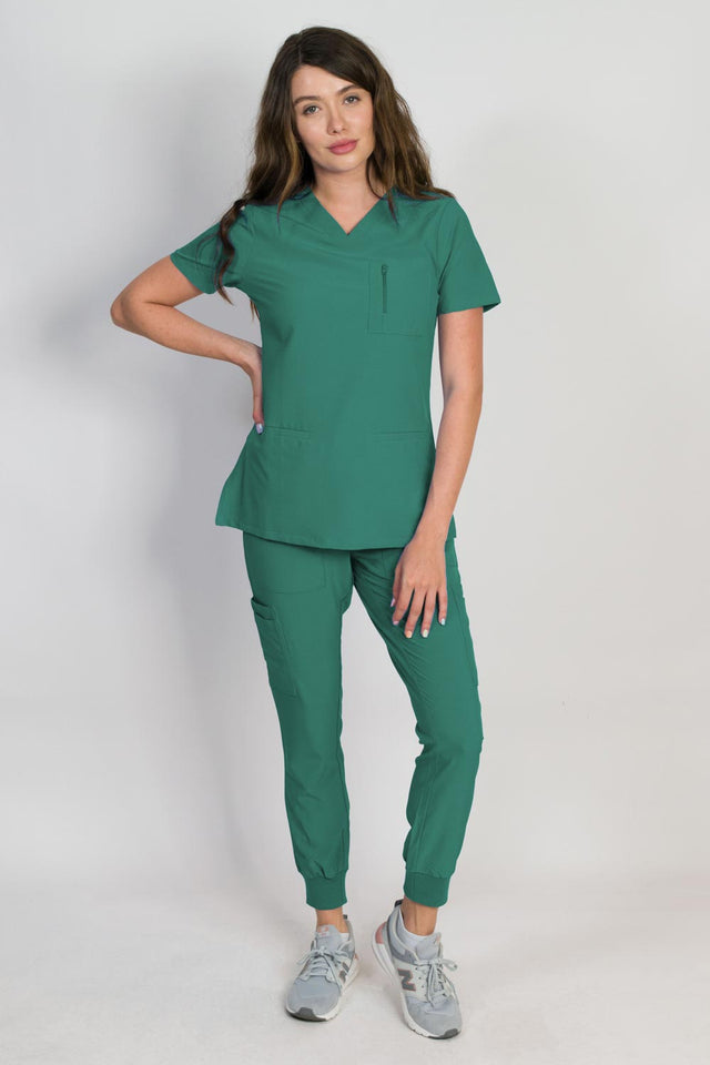 A female model wearing a hunter green scrub set with mitered neck zip chest pocket top and matching jogger pants with multiple pockets, designed for medical professionals.