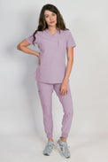 A female model wearing a lavender scrub set with mitered neck zip chest pocket top and matching jogger pants with multiple pockets, designed for medical professionals.