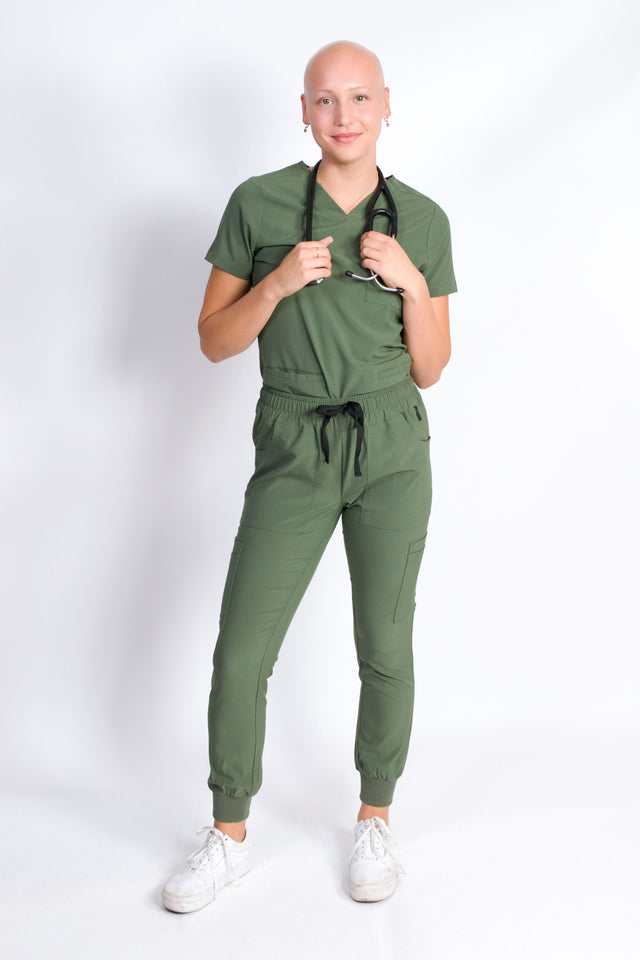 A female model wearing an olive green scrub set with mitered neck zip chest pocket top and matching jogger pants with multiple pockets, designed for medical professionals.