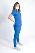 A female model wearing a royal blue scrub set with mitered neck zip chest pocket top and matching jogger pants with multiple pockets, designed for medical professionals.