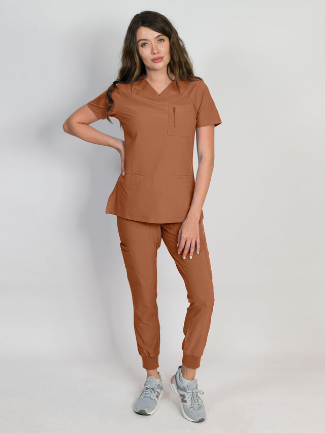 Terracotta brown scrub set with a mitered neck V-neck top and jogger pants, both with multiple pockets, made from a smooth stretch fabric.