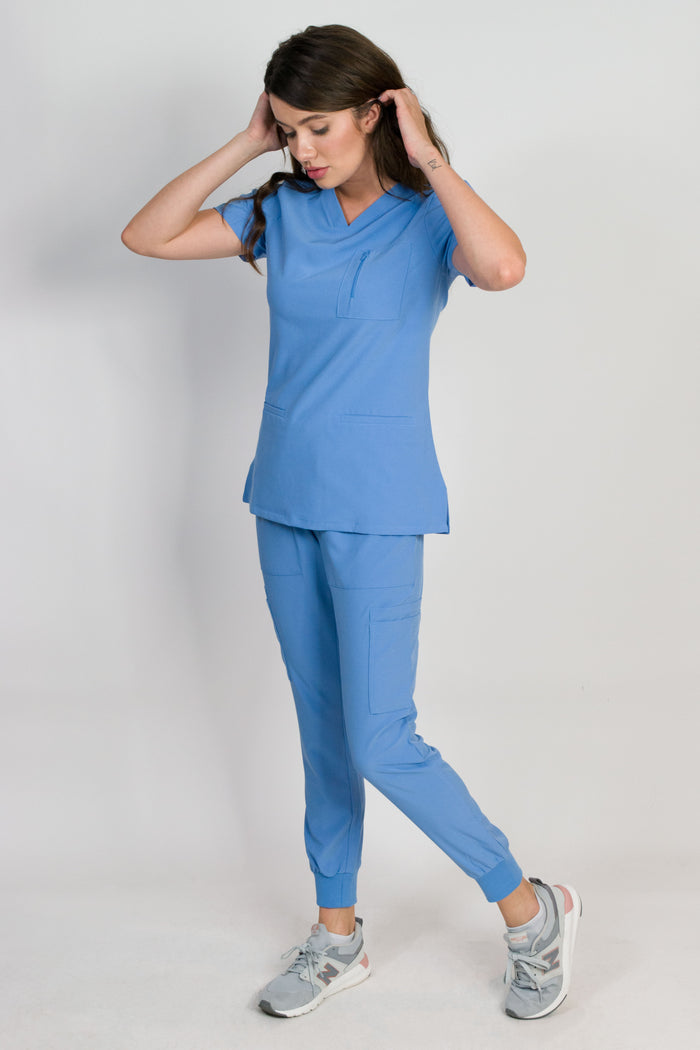 A female model wearing a ceil blue scrub set with mitered neck zip chest pocket top and matching jogger pants with multiple pockets, designed for medical professionals.
