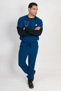 A man wearing a blue V-neck scrub set with 4 pockets and rib knit cuffs. The set includes a top with a chest patch pocket and bottom hem pockets, along with matching pants with inset trouser pockets and back welt pockets.