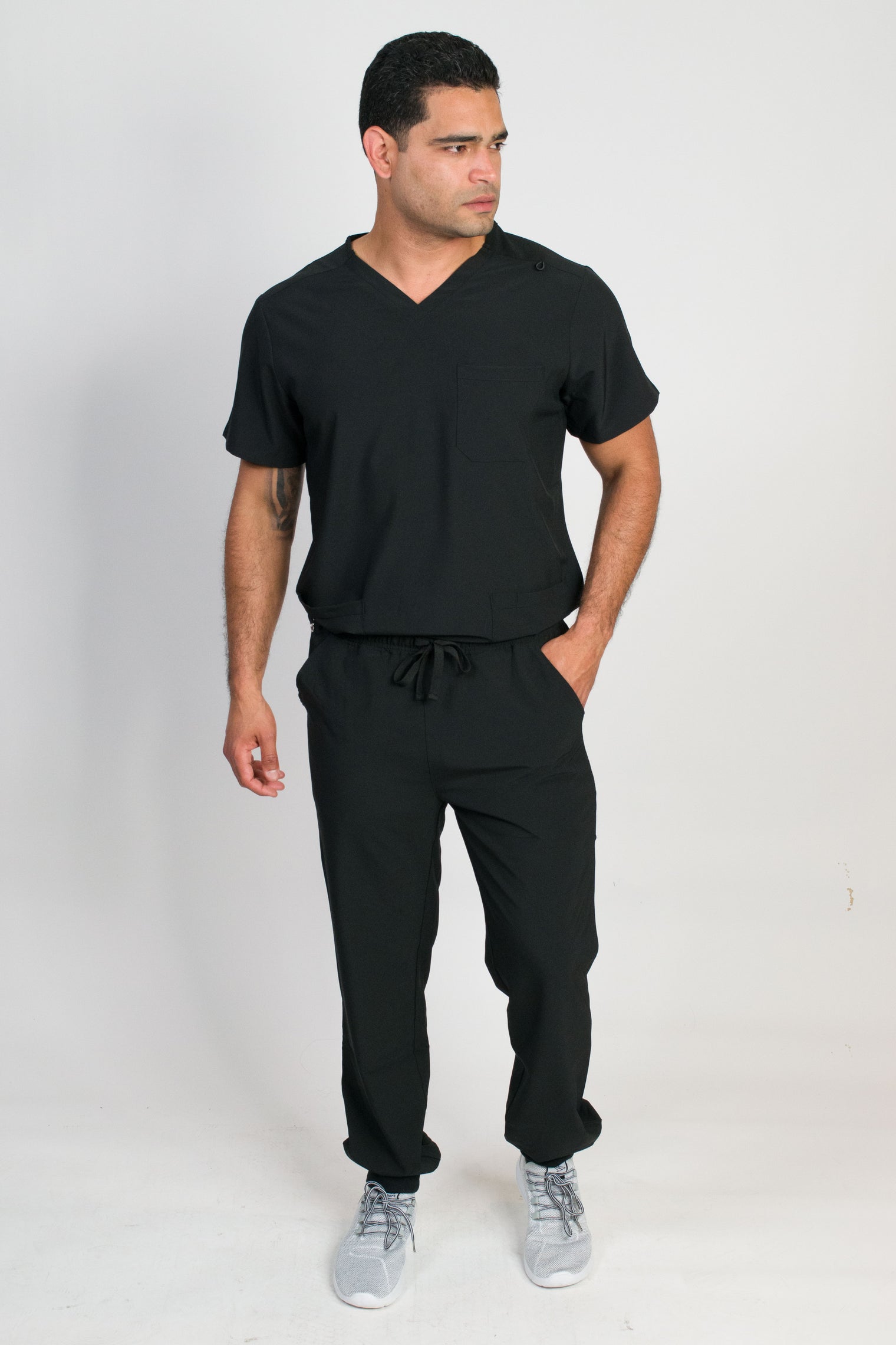 A man wearing a black V-neck scrub set with 4 pockets and rib knit cuffs. The set includes a top with a chest patch pocket and bottom hem pockets, along with matching pants with inset trouser pockets and back welt pockets.